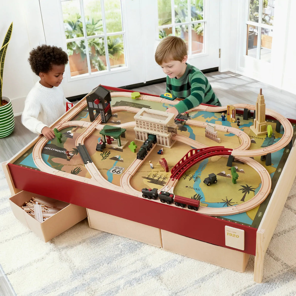 Two young children play with the Tiny Land® 1920 Railway Adventure Train Table by Tiny Land INC, featuring tracks, buildings, a red bridge, and toy vehicles on a large play table in a bright room with windows and plants outside.
