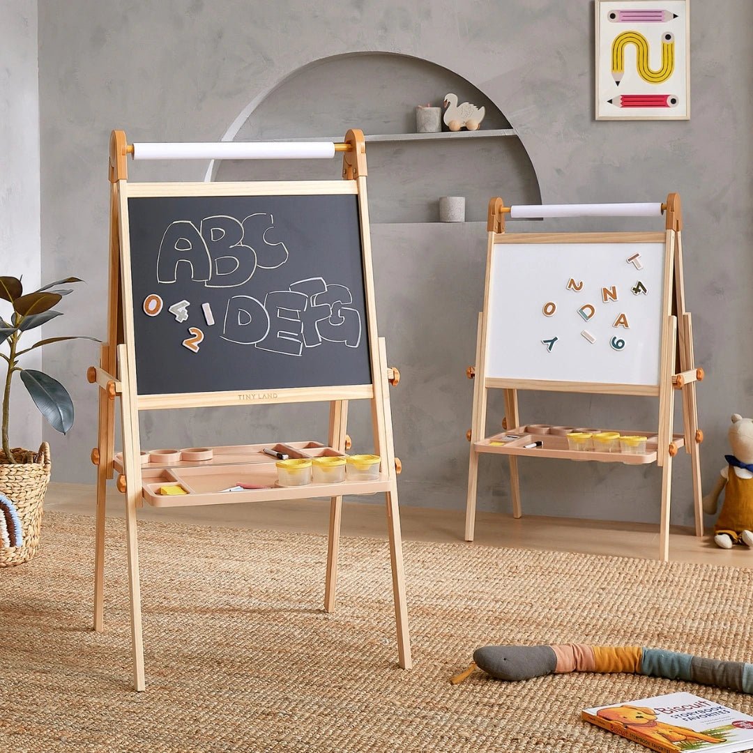 A playroom with two wooden kids art easels: one is the Tiny Land® Magnitales Easel for Kids by Tiny Land, featuring magnetic letters; the other is a blackboard with ABC and doodles. Art supplies and toys are scattered on a woven rug and shelves.