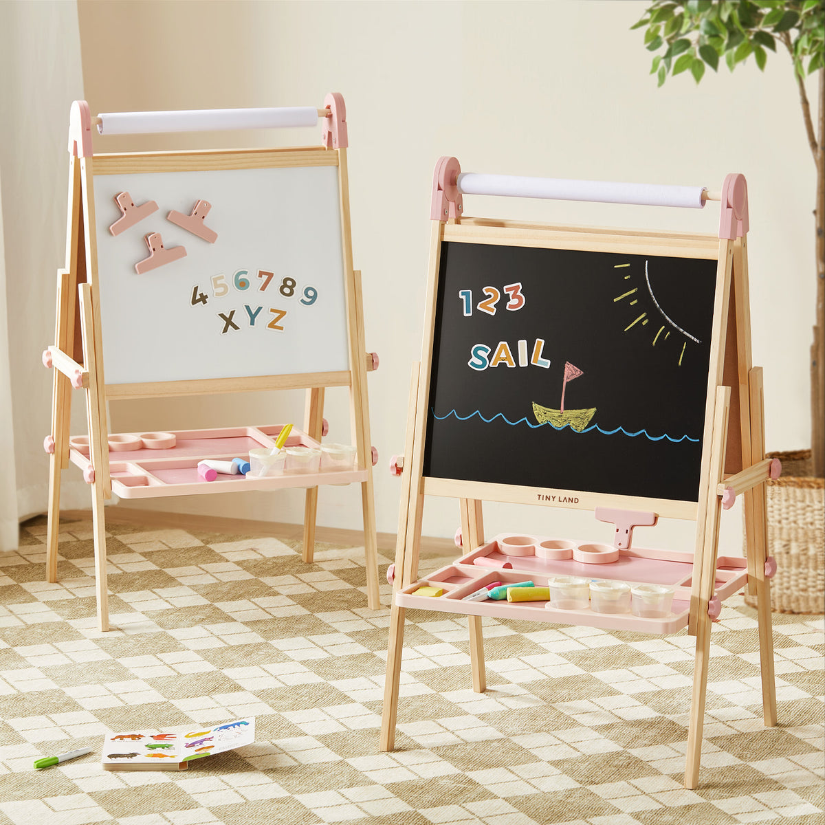 Two Tiny Land® Magnitales Easel for Kids - Pink by Tiny Land stand on a checkered rug: one displays magnetic letters and numbers, the other shows “123 SAIL” with a sailboat. Art supplies sit on trays beneath each easel.