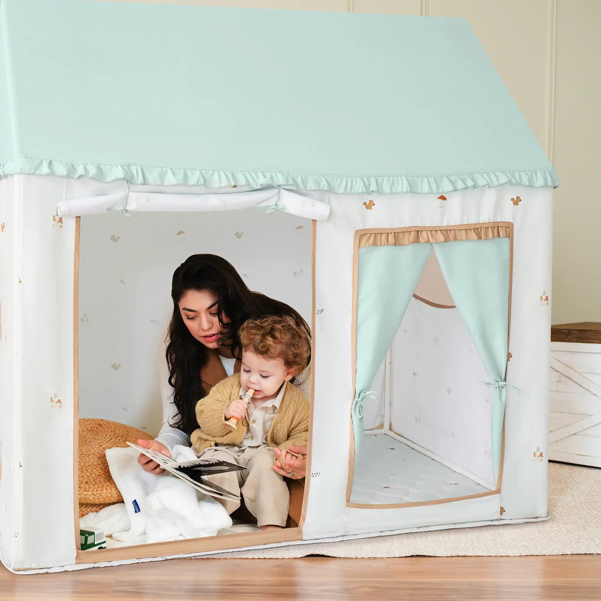 A woman reads a book to a young boy holding a stuffed animal inside the Tiny Land® Sweet Woodland Kids Play Tent by Tiny Land INC, a cozy white playhouse with light blue roof and curtains—perfect for storytime.