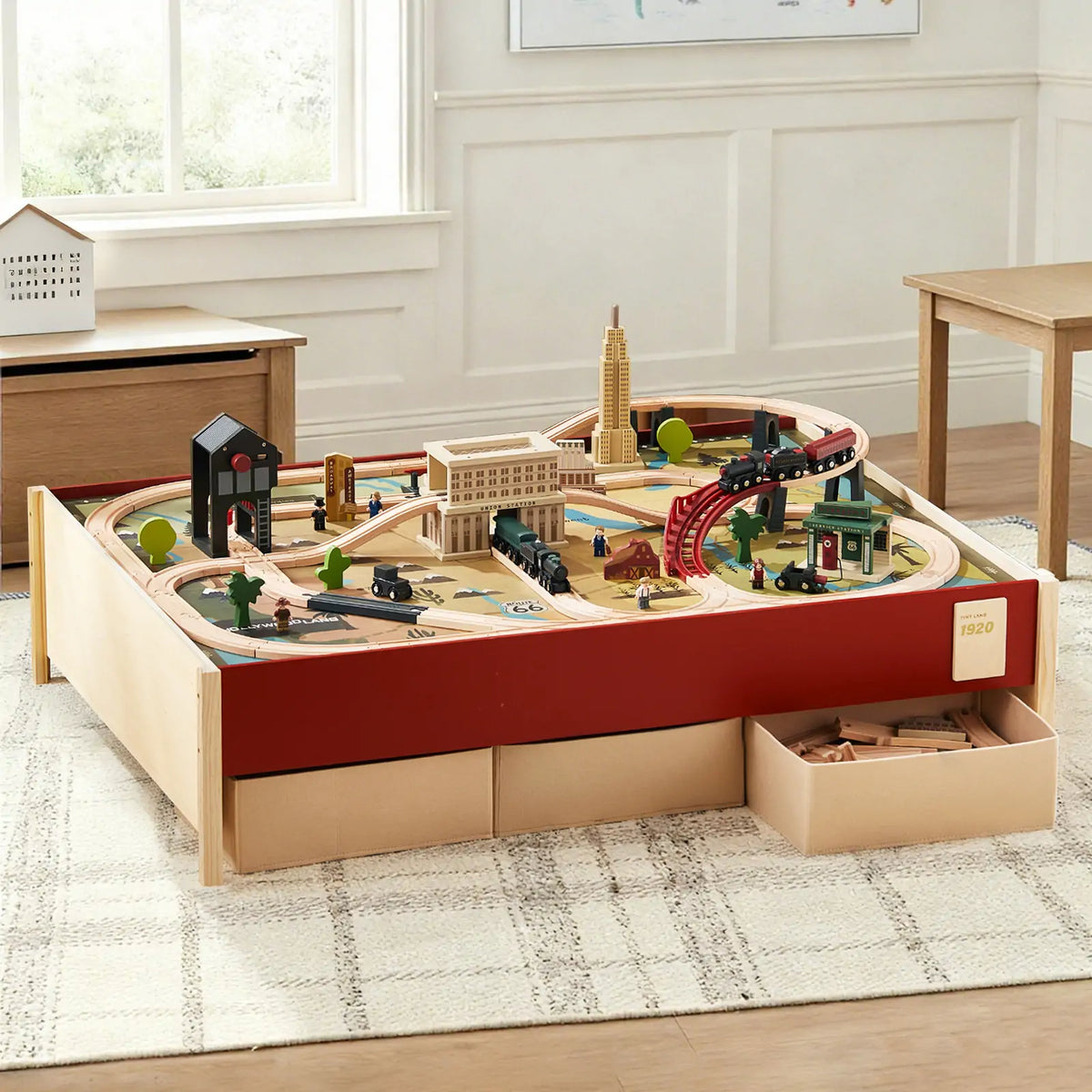 The Tiny Land® 1920 Railway Adventure Train Table by Tiny Land INC features an interactive city playset with tracks, bridges, and buildings. Two drawers store extra pieces, while the table sits on a patterned rug in a bright, tidy room.