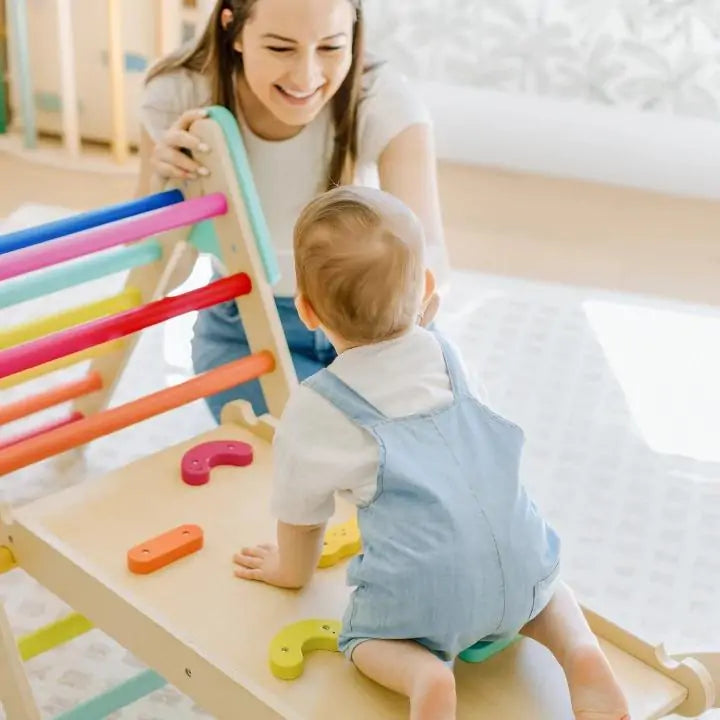 A smiling woman watches a baby in blue overalls climbing on the Tiny Land® 7-in-1 Rainbow Pikler Triangle Set by Tiny Land, featuring colorful bars and a reversible ramp in a bright, cheerful room.