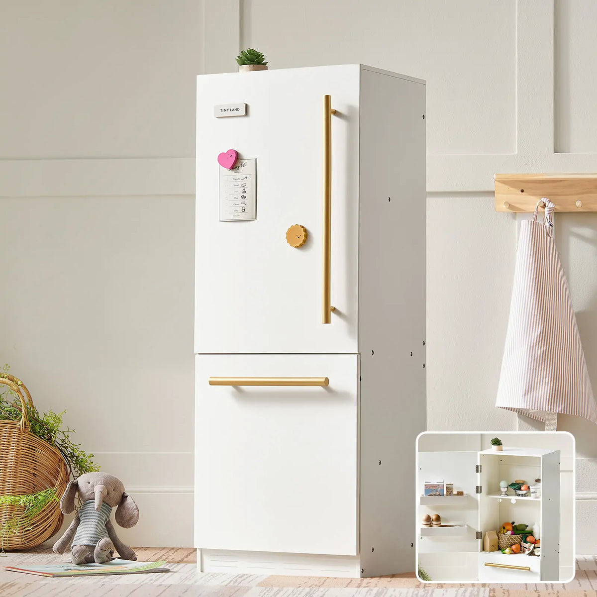 The Tiny Land® Play Kitchen Fridge, a white wooden refrigerator with gold handles, stands in a bright room. A note and heart magnet are on the door, a stuffed elephant sits nearby, and an inset shows toy food inside—ideal for any play kitchen.