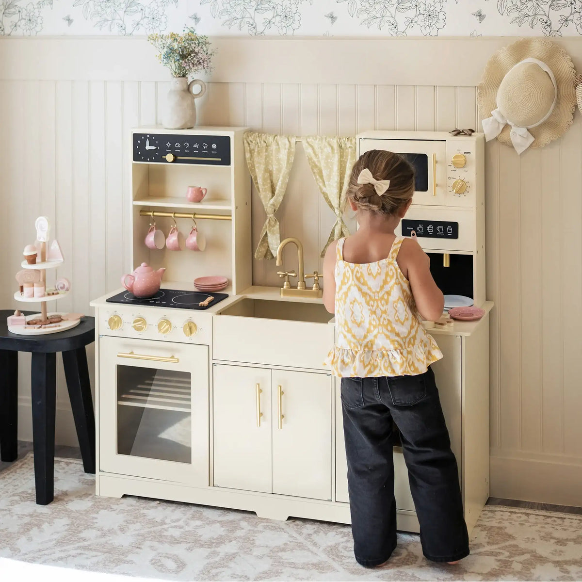 A young child with a hair bow plays in a cozy, well-decorated room at the Tiny Land® Iconic Wooden Play Kitchen—Cream Pretend Cooking Set for Kids—featuring gold details, pink dishes, and patterned curtains.
