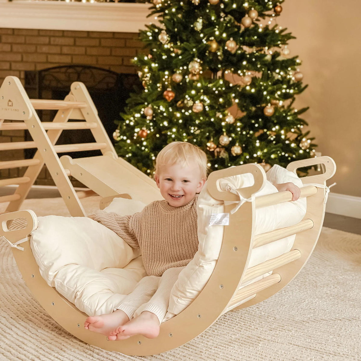 A smiling blonde child sits on a cushioned wooden rocker in a cozy room, feet resting on the Tiny Land® Thick Padded Play Cushion, with a festive tree behind. The barefoot child wears a beige sweater and light pants on a soft rug. Arch not included.