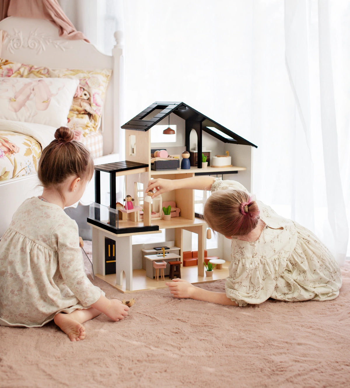 Two children sit on a pink carpet, playing with the Tiny Land® Sweetwood Edge Dollhouse by Tiny Land and arranging miniature family figures in its cozy, sunlit bedroom.