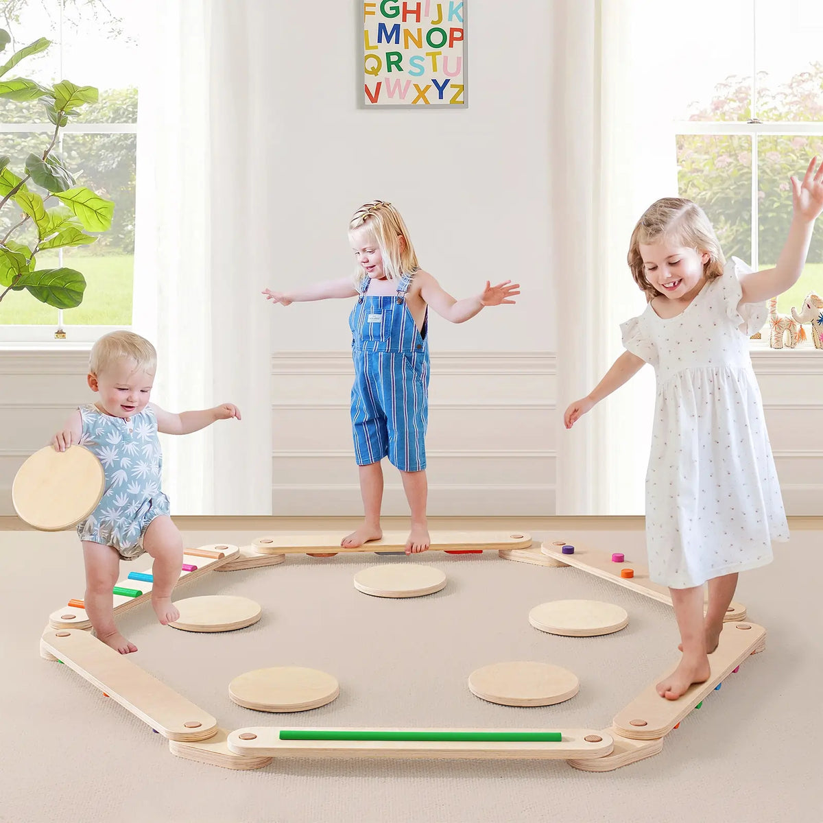 Three young children play and balance on the Tiny Land® Balance Beam by Tiny Land in a bright, sunlit room with a soft carpet. A colorful alphabet poster hangs on the wall behind them.