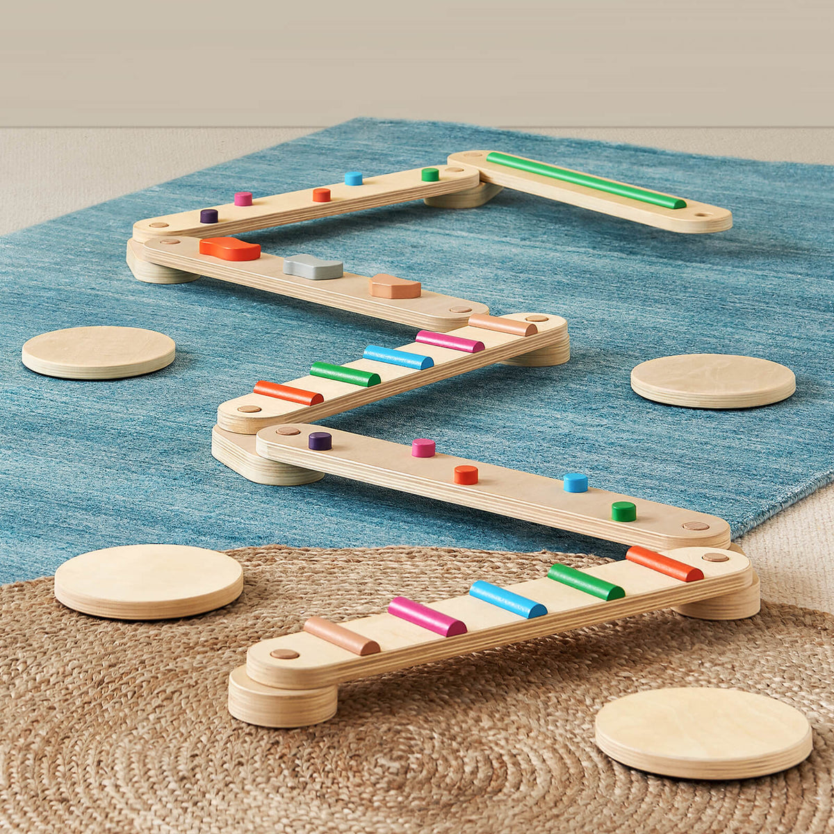 The Tiny Land® Balance Beam by Tiny Land features wooden beams with colorful cylindrical pegs in a zigzag on a blue rug and mat, creating a fun kids’ coordination toy, with round wooden stepping platforms placed nearby.