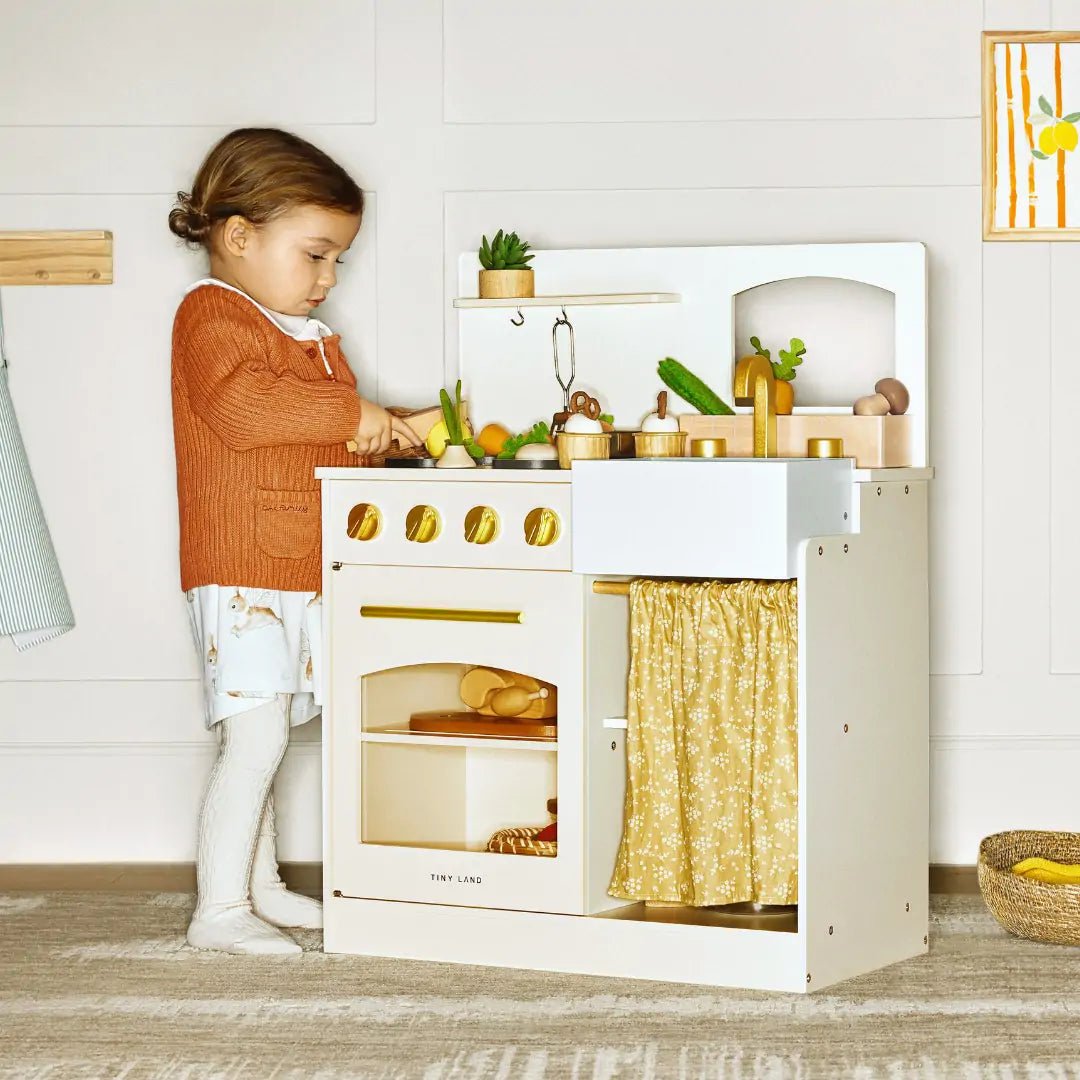 A young child in a rust sweater and white tights enjoys imaginative play with the Tiny Land® Wooden Play Kitchen for Toddlers - Honey Kitchenette Pretend Cooking Set by Tiny Land in a cozy, warmly lit room.
