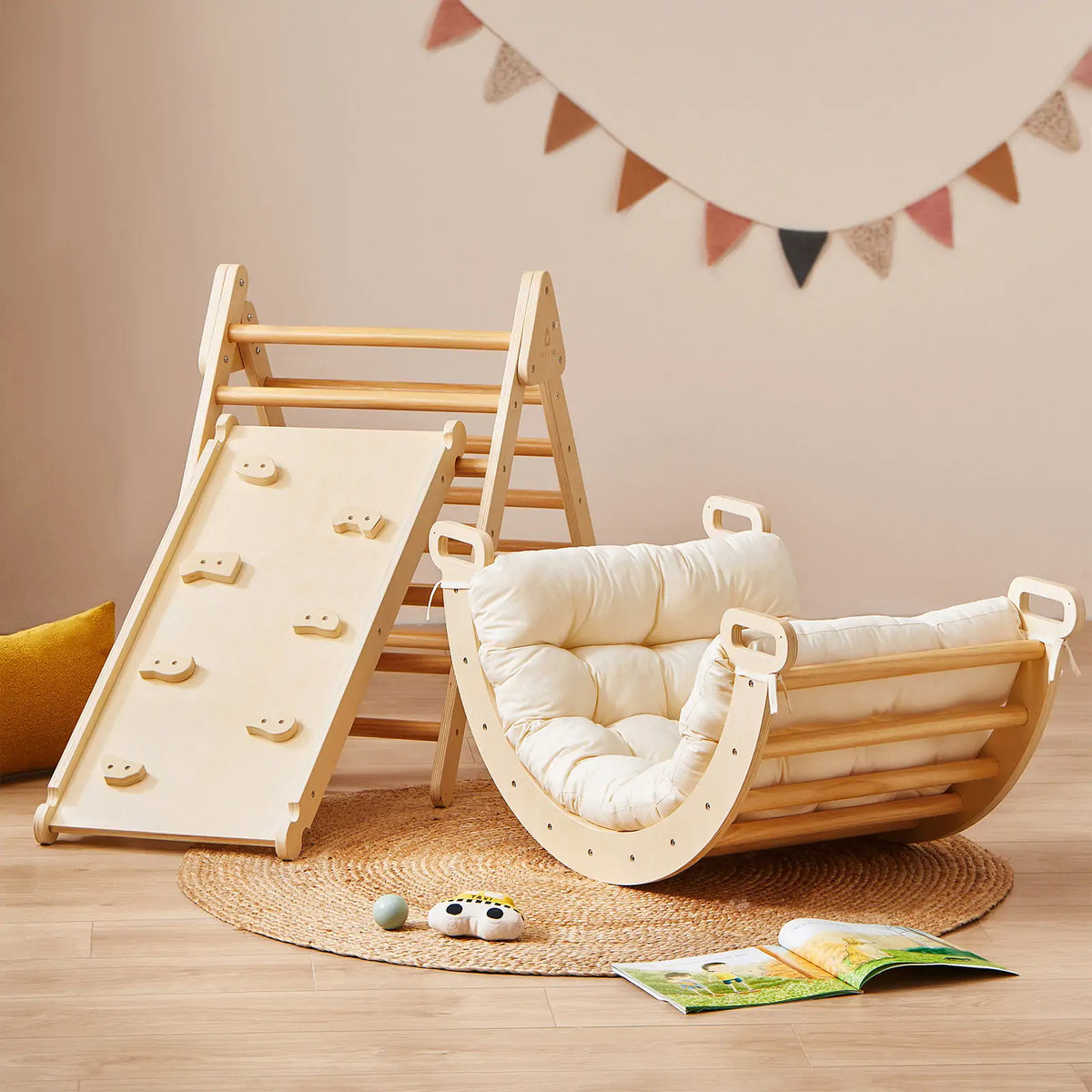 The Tiny Land® 7-in-1 Montessori Climbing Set with Play Pad by Tiny Land, featuring a ramp, ladder, and cushioned rocker, is set on a round woven rug. Nearby sit a yellow pillow, padded cushion, soft toy, open book, and colorful wall bunting.