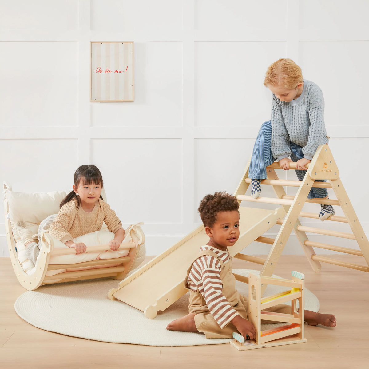 Three young children play indoors with the Tiny Land® 7-in-1 Montessori Climbing Set with Play Pad: one girl rocks, a boy climbs the Pikler triangle and slide, and another explores a wooden ramp on a rug beside the colorful padded play pad.