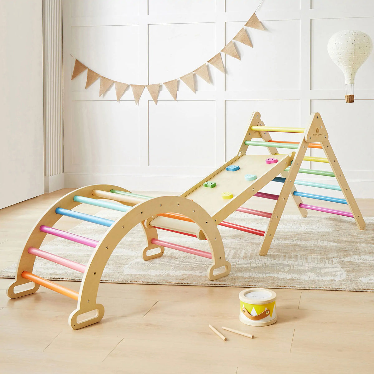 The Tiny Land® 7-in-1 Rainbow Climbing Set by Tiny Land includes a Pikler Triangle, wooden arch ladder, and slanted climbing board with colorful grips. A toy drum and stick also sit on the light wood floor in this bright room.