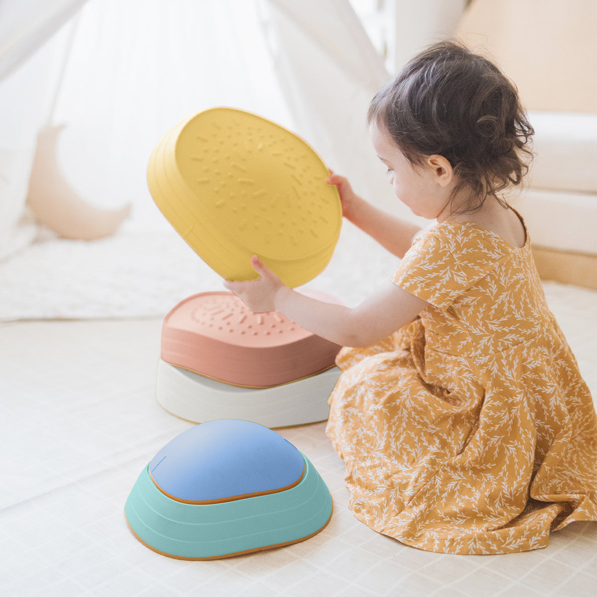 A young child in a yellow patterned dress sits on the floor of a cozy room, stacking colorful Tiny Land® Stepping Stones - Beach Vacation, building motor skills through fun obstacle play with these textured stones by Tiny Land.