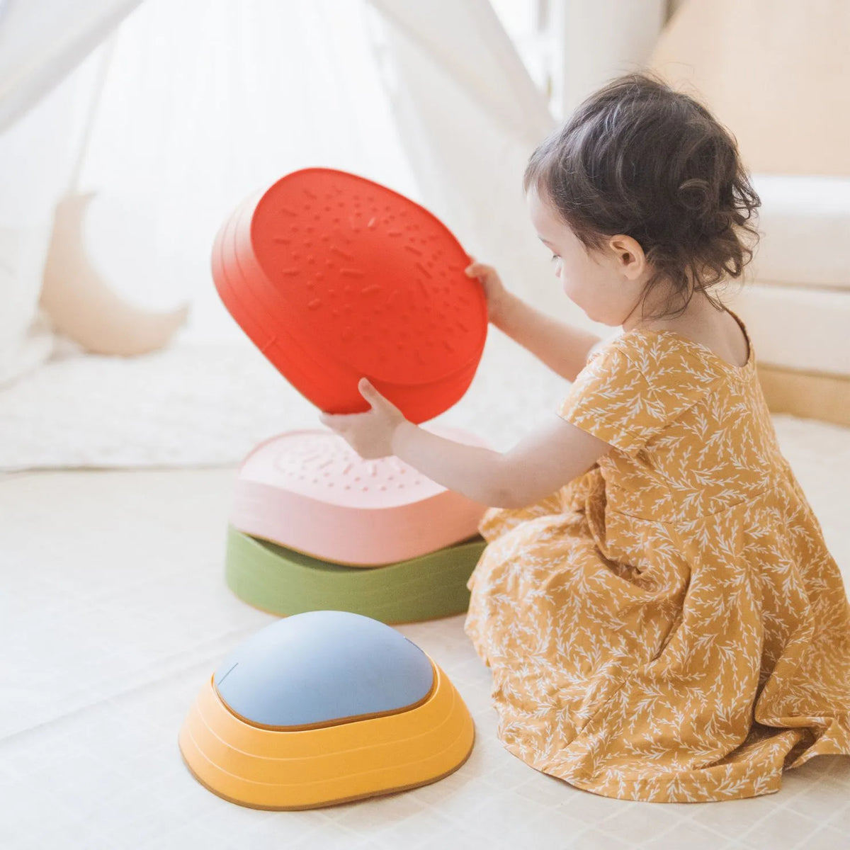 A young child in a yellow dress sits on the floor, stacking Tiny Land® Stepping Stones - Vintage Color by Tiny Land in a bright, cozy room—building motor skills for a playful obstacle course.