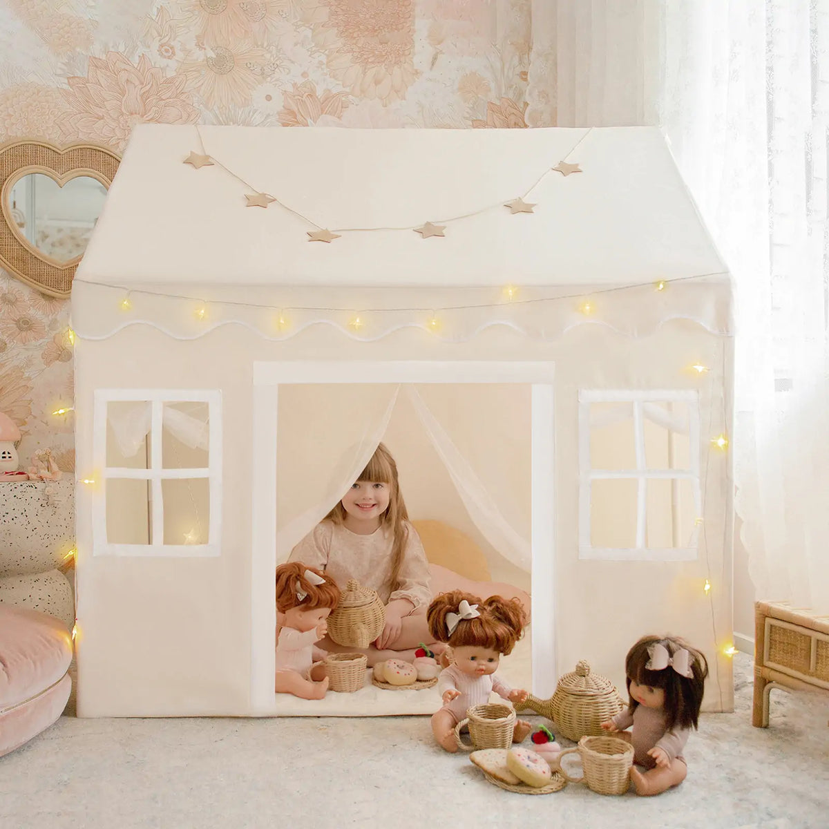 A young girl enjoys the Tiny Land® Playhouse Tent – Natural Cream by Tiny Land, decorated with string lights and surrounded by dolls, tea sets, and baskets as soft natural light streams in through a window with floral wallpaper.