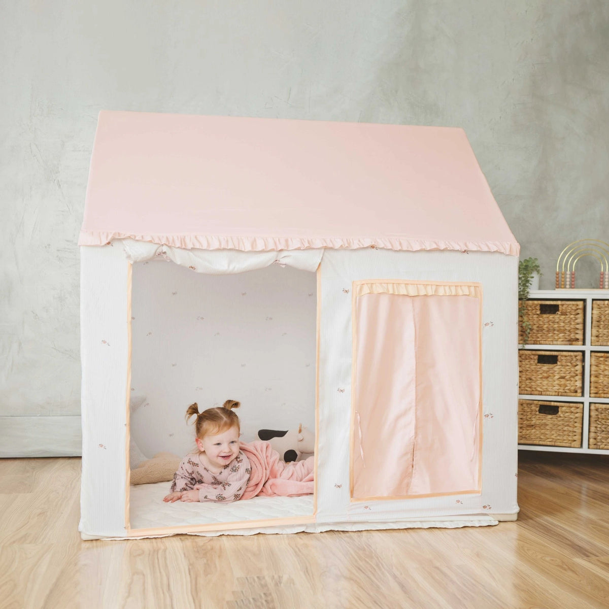 A young child in pink pajamas smiles while lying inside the Tiny Land® Sweet Citrus Kids Play Tent – a white and pink indoor playhouse by Tiny Land, set up on a wooden floor in a cozy, modern room.