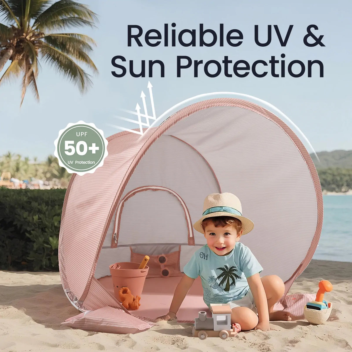 A young child plays with toys on the sand inside the Tiny Land® Baby Beach Tent – Blush Pink, which offers UPF 50+ UV protection. The tent by Tiny Land is set against a scenic backdrop of palm trees and mountains.