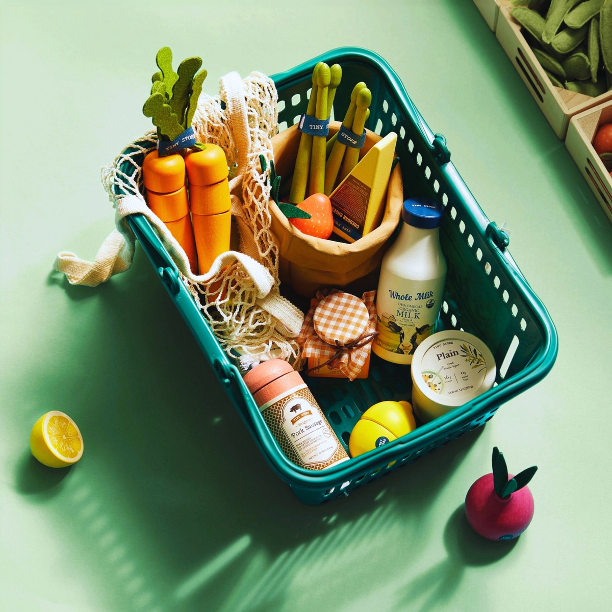 The Tiny Land® Organic Play Food Collection by Tiny Land INC features a green shopping basket with plush and wooden groceries like carrots, asparagus, milk, a jar, can, lemon half, and apple displayed on a light green surface.